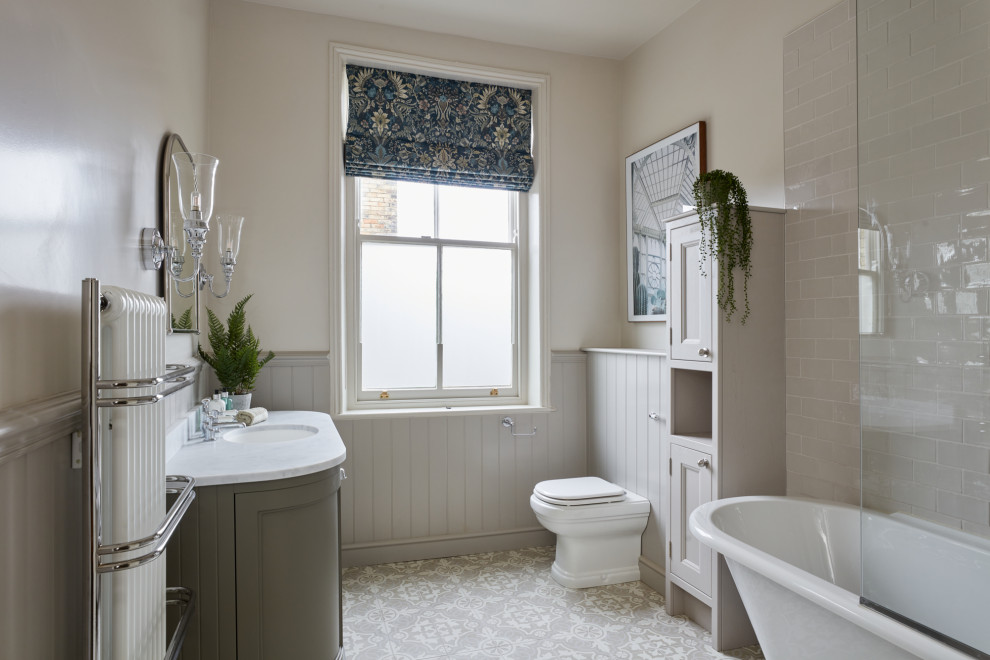 This is an example of a country bathroom in Other with recessed-panel cabinets, grey cabinets, a claw-foot bath, a shower/bath combination, a one-piece toilet, beige walls, a submerged sink, grey floors, an open shower, white worktops, a single sink, a freestanding vanity unit and wainscoting.