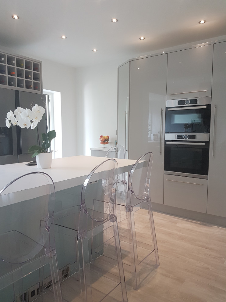 This is an example of a medium sized modern l-shaped kitchen/diner in West Midlands with a double-bowl sink, flat-panel cabinets, grey cabinets, quartz worktops, blue splashback, glass sheet splashback, black appliances, medium hardwood flooring, an island, brown floors and white worktops.