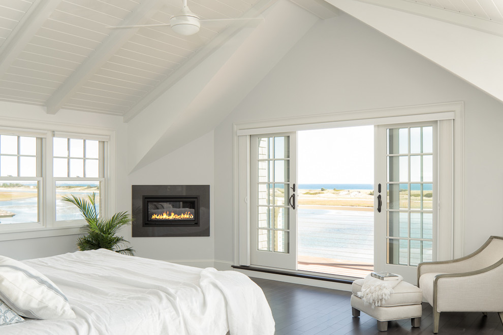 This is an example of a large beach style master bedroom in Portland Maine with white walls, dark hardwood floors, a ribbon fireplace and a tile fireplace surround.