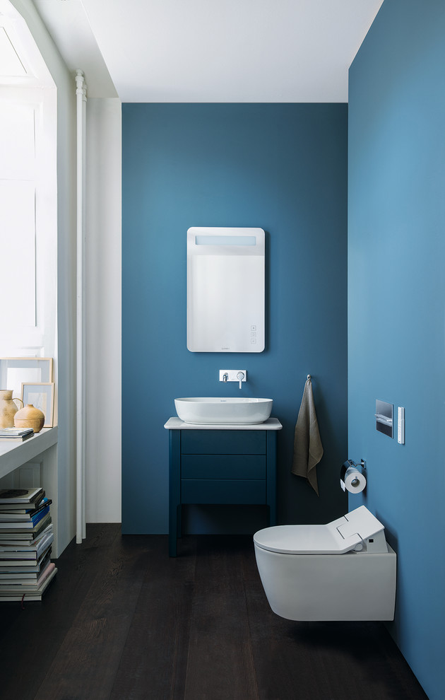 This is an example of a small contemporary bathroom with blue cabinets, a vessel sink, engineered quartz benchtops, flat-panel cabinets, a wall-mount toilet, blue walls, dark hardwood floors, brown floor and white benchtops.