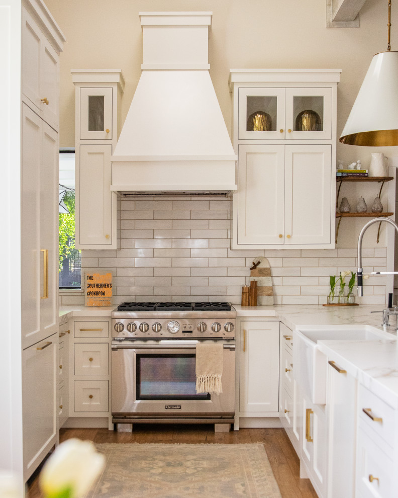 This is an example of a country u-shaped kitchen in San Francisco with a farmhouse sink, shaker cabinets, white cabinets, white splashback, stainless steel appliances, medium hardwood floors, a peninsula, brown floor and white benchtop.