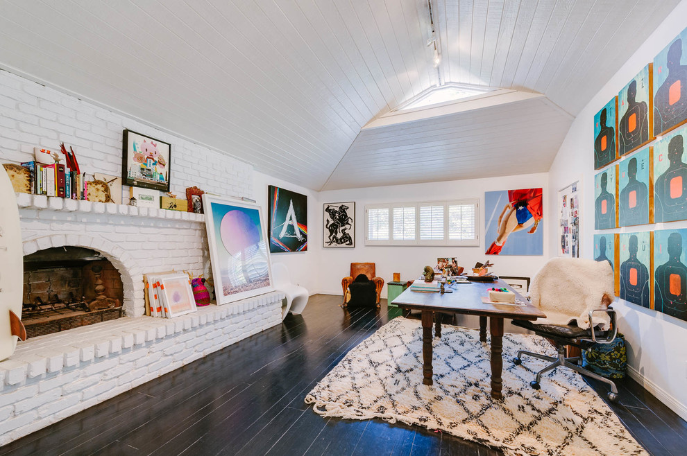 This is an example of an expansive eclectic home studio in Los Angeles with white walls, a standard fireplace, a brick fireplace surround, a freestanding desk, black floor and dark hardwood floors.