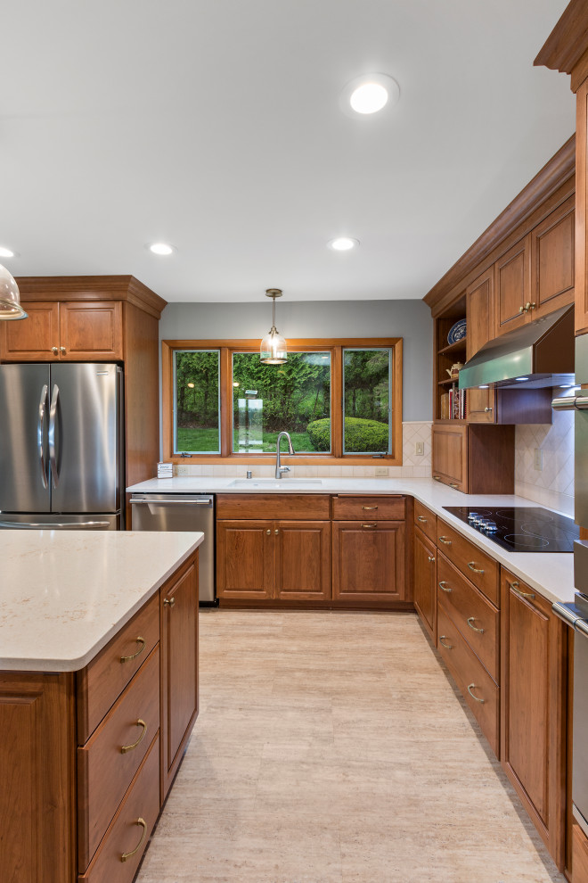 Photo of a mid-sized traditional l-shaped eat-in kitchen in Milwaukee with an undermount sink, raised-panel cabinets, brown cabinets, quartz benchtops, beige splashback, stone tile splashback, stainless steel appliances, vinyl floors, with island, beige floor and white benchtop.