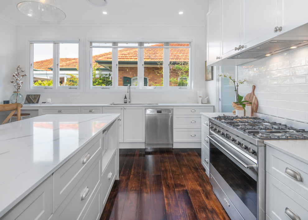 Photo of a large transitional l-shaped open plan kitchen in Brisbane with a double-bowl sink, shaker cabinets, grey cabinets, solid surface benchtops, white splashback, subway tile splashback, stainless steel appliances, dark hardwood floors, with island and white benchtop.