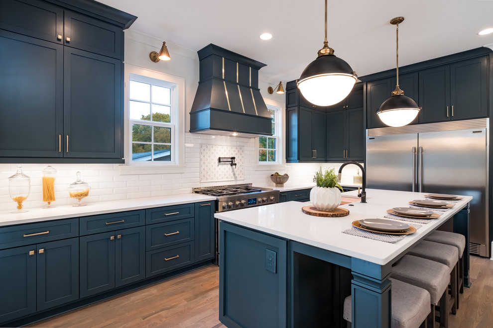 This is an example of a country l-shaped kitchen in Charlotte with an undermount sink, shaker cabinets, blue cabinets, white splashback, subway tile splashback, stainless steel appliances, medium hardwood floors, with island, brown floor and white benchtop.