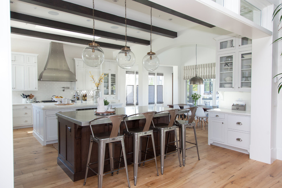 Inspiration for a large transitional l-shaped light wood floor eat-in kitchen remodel in Phoenix with a farmhouse sink, white backsplash, stainless steel appliances, an island, shaker cabinets, white cabinets, stainless steel countertops and porcelain backsplash