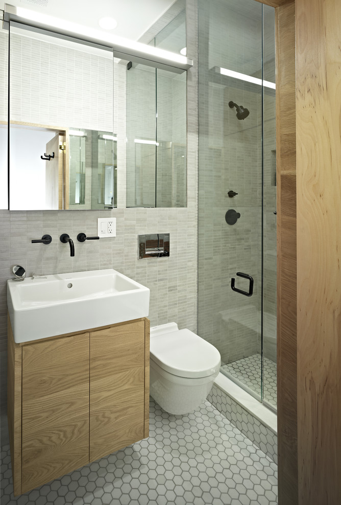This is an example of a contemporary bathroom in New York with a wall-mount sink, flat-panel cabinets, an alcove shower, a wall-mount toilet, gray tile and light wood cabinets.