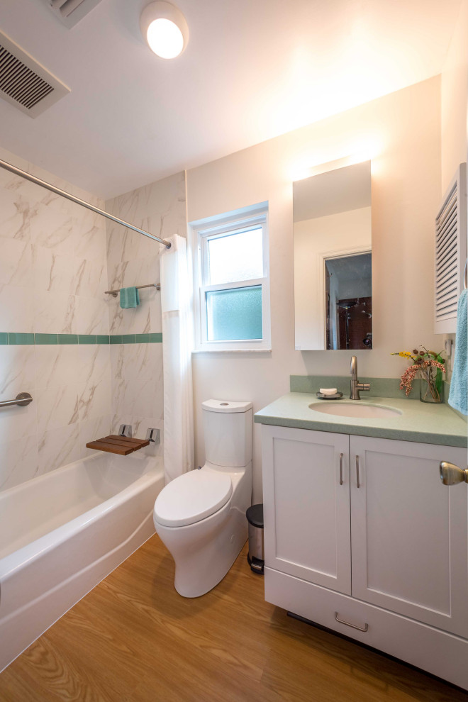 This is an example of a mid-sized traditional kids bathroom with recessed-panel cabinets, white cabinets, an alcove tub, an alcove shower, a one-piece toilet, white tile, marble, beige walls, light hardwood floors, a drop-in sink, solid surface benchtops, brown floor, a shower curtain and green benchtops.