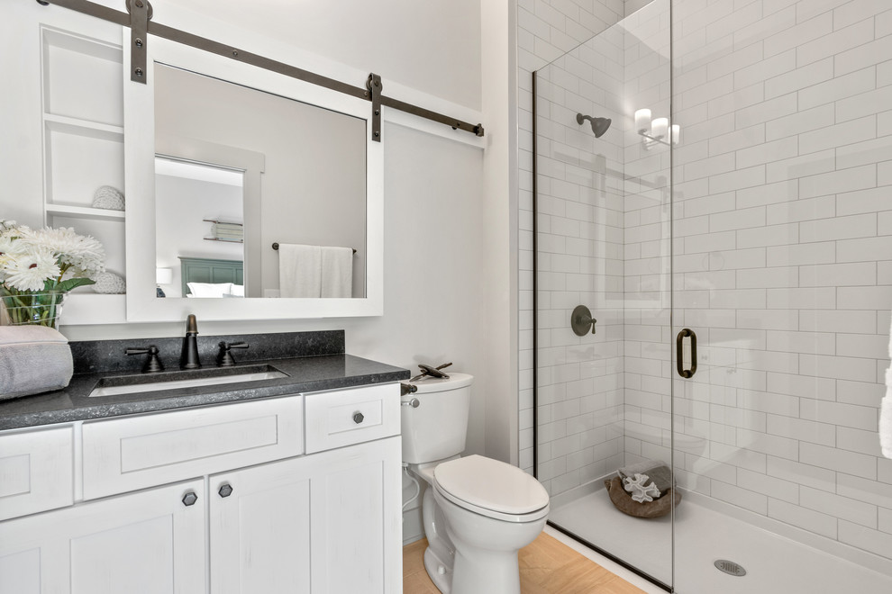 Inspiration for a country bathroom in Columbus with shaker cabinets, white cabinets, a curbless shower, a two-piece toilet, white tile, subway tile, white walls, medium hardwood floors, an undermount sink, brown floor, a hinged shower door and black benchtops.