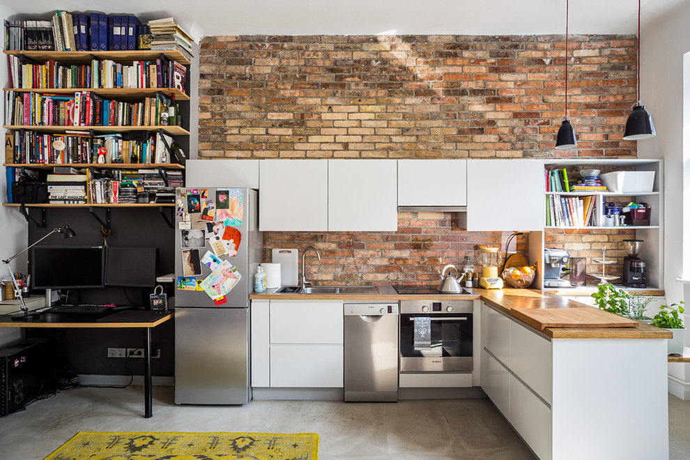 Photo of a small industrial l-shaped kitchen in Other with flat-panel cabinets, white cabinets, wood benchtops, red splashback, stainless steel appliances, concrete floors and a peninsula.