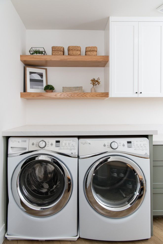 Photo of a mid-sized beach style dedicated laundry room in Orange County with a drop-in sink, green cabinets, quartz benchtops, white splashback, engineered quartz splashback, white walls, medium hardwood floors, a side-by-side washer and dryer and white benchtop.
