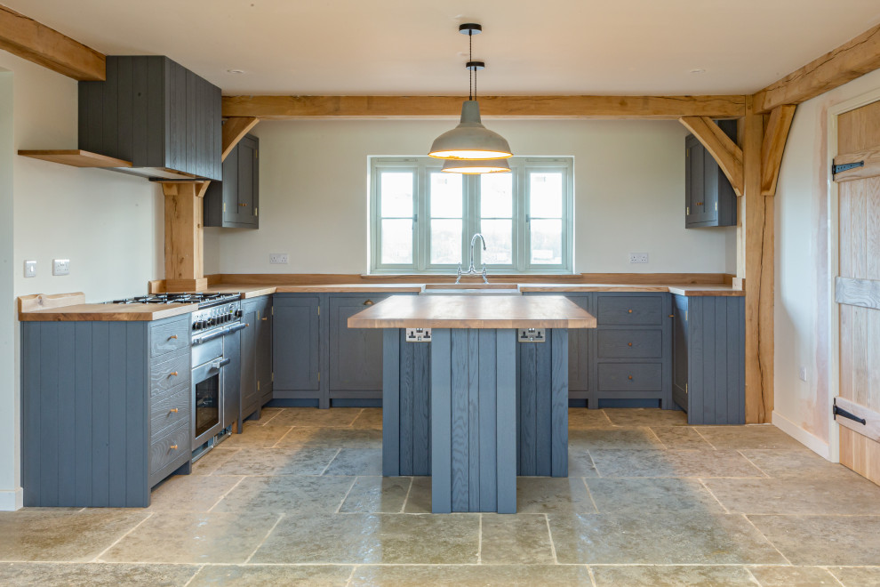 This is an example of a mid-sized country u-shaped eat-in kitchen in West Midlands with a farmhouse sink, shaker cabinets, grey cabinets, wood benchtops, stainless steel appliances, travertine floors, with island, grey floor and brown benchtop.