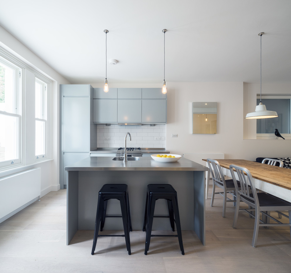Photo of a small contemporary galley eat-in kitchen in London with an integrated sink, flat-panel cabinets, grey cabinets, stainless steel benchtops, white splashback, subway tile splashback, light hardwood floors and with island.