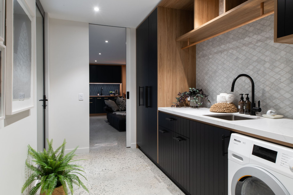 This is an example of a contemporary single-wall laundry room in Hobart with an undermount sink, flat-panel cabinets, black cabinets, grey splashback, mosaic tile splashback, white walls, grey floor and white benchtop.