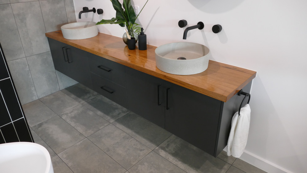 This is an example of a large industrial master bathroom in Other with black cabinets, a freestanding tub, gray tile, ceramic tile, white walls, ceramic floors, a vessel sink, wood benchtops, grey floor, orange benchtops, a double vanity and a floating vanity.
