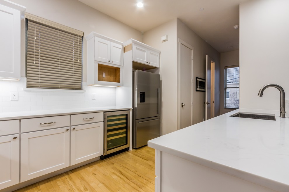 Small traditional u-shaped kitchen in Dallas with flat-panel cabinets, white cabinets, quartz benchtops, white splashback, subway tile splashback, stainless steel appliances, light hardwood floors, with island and white benchtop.