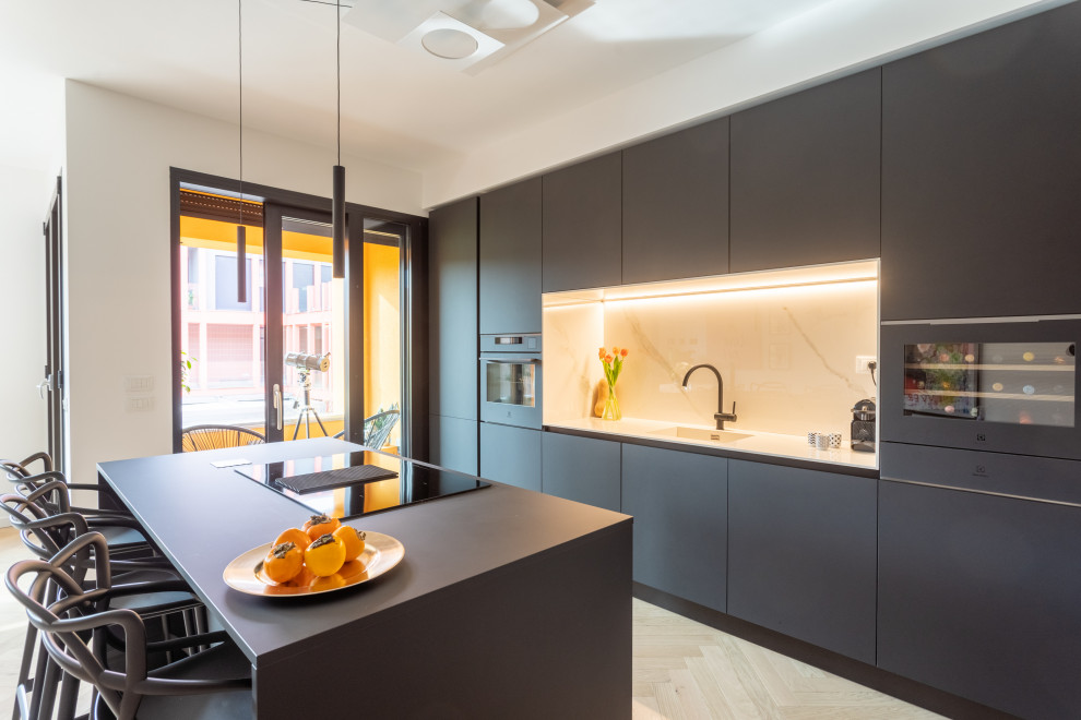 Design ideas for a mid-sized modern galley open plan kitchen in Milan with a drop-in sink, flat-panel cabinets, black cabinets, tile benchtops, white splashback, porcelain splashback, black appliances, light hardwood floors, with island, brown floor and white benchtop.