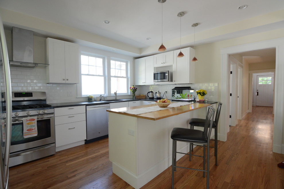 Inspiration for a large transitional l-shaped medium tone wood floor and brown floor kitchen pantry remodel in Boston with a drop-in sink, flat-panel cabinets, white cabinets, solid surface countertops, white backsplash, subway tile backsplash, stainless steel appliances and an island