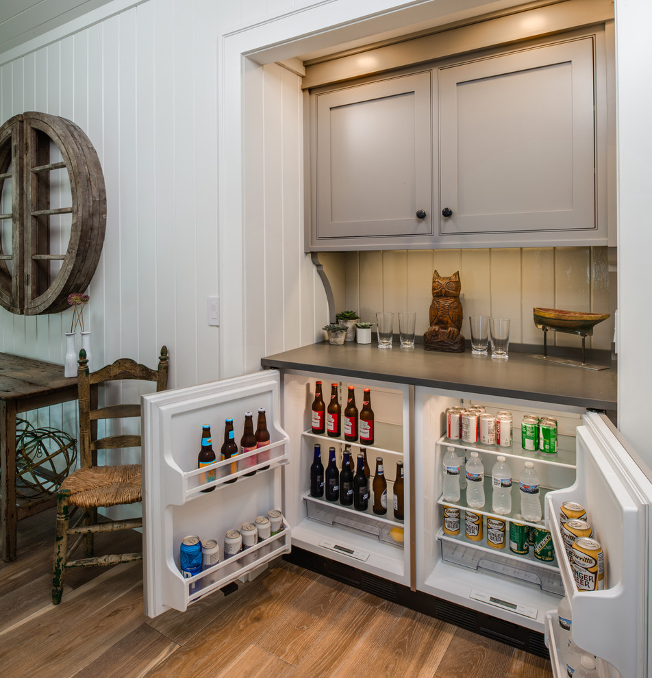 This is an example of a small beach style single-wall wet bar in Other with no sink, white splashback, timber splashback, medium hardwood floors, brown floor, grey benchtop, recessed-panel cabinets, grey cabinets and quartz benchtops.
