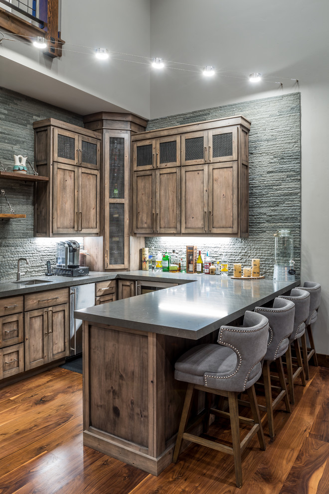 Photo of a mid-sized country u-shaped seated home bar in Sacramento with an undermount sink, shaker cabinets, medium wood cabinets, solid surface benchtops, grey splashback, stone slab splashback, dark hardwood floors and brown floor.