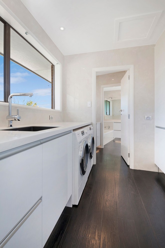 Mid-sized contemporary galley dedicated laundry room in Christchurch with an undermount sink, white cabinets, solid surface benchtops, vinyl floors and a side-by-side washer and dryer.