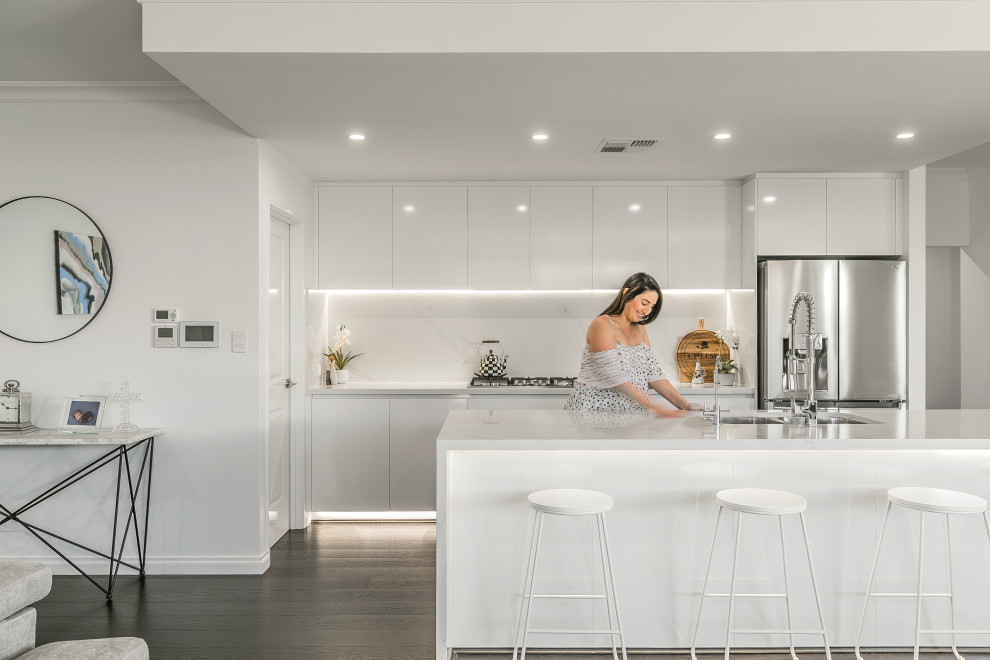 Contemporary open plan kitchen in Perth with a double-bowl sink, flat-panel cabinets, white cabinets, white splashback, stainless steel appliances, with island, brown floor and white benchtop.
