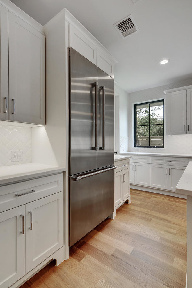 Photo of a modern kitchen in Austin with shaker cabinets, white cabinets, quartz benchtops, white splashback, ceramic splashback, stainless steel appliances and light hardwood floors.