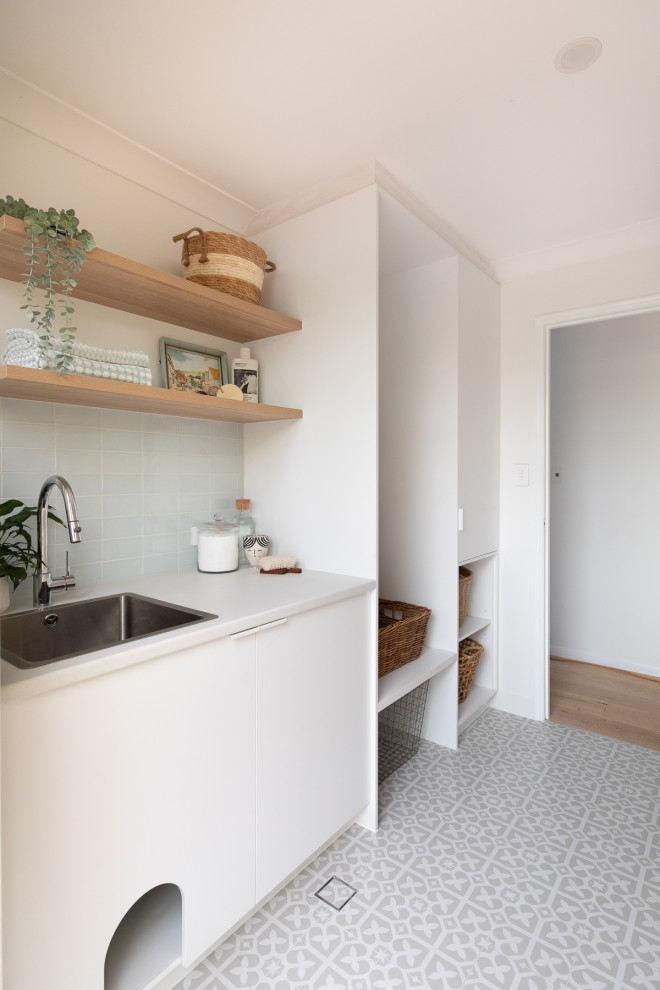 Design ideas for a mid-sized beach style single-wall dedicated laundry room in Brisbane with a drop-in sink, flat-panel cabinets, white cabinets, laminate benchtops, blue splashback, porcelain splashback, white walls, porcelain floors, a stacked washer and dryer, grey floor and white benchtop.