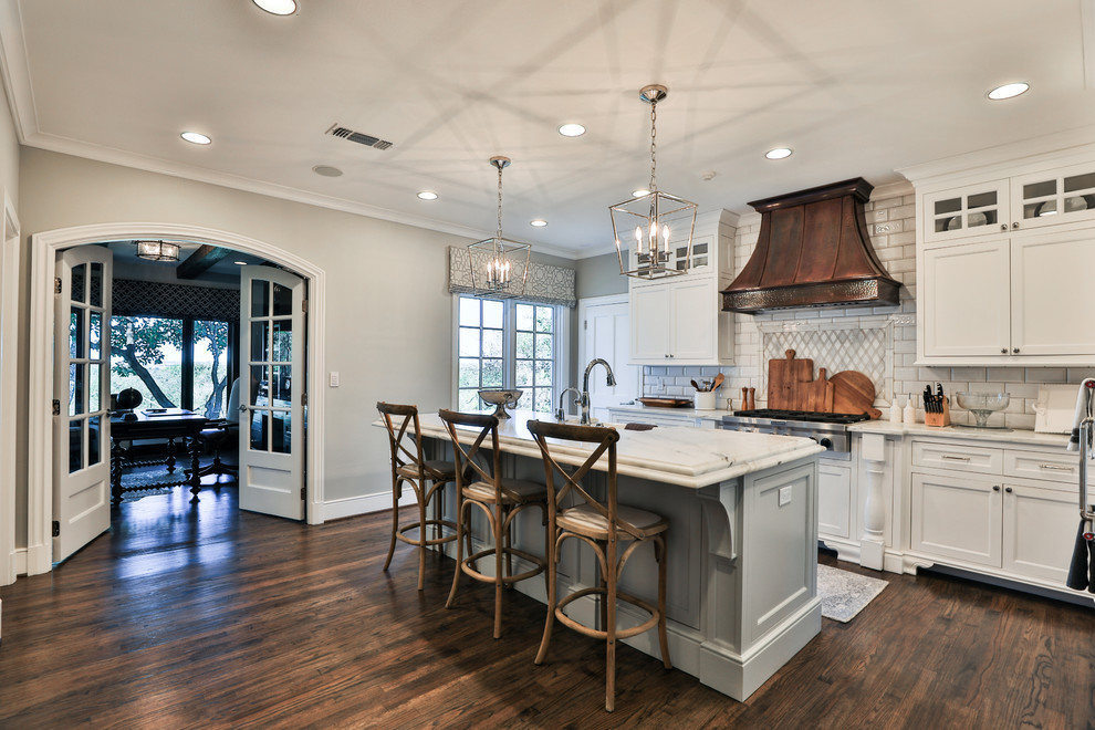 Inspiration for a mid-sized transitional l-shaped medium tone wood floor and brown floor kitchen remodel in Dallas with a farmhouse sink, recessed-panel cabinets, white cabinets, marble countertops, white backsplash, subway tile backsplash, paneled appliances and an island