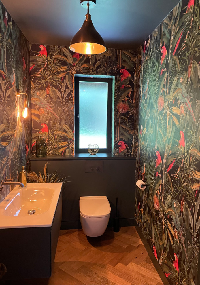 Small contemporary powder room in Hertfordshire with grey cabinets, a wall-mount toilet, multi-coloured walls, light hardwood floors, a wall-mount sink, brown floor, a floating vanity and wallpaper.