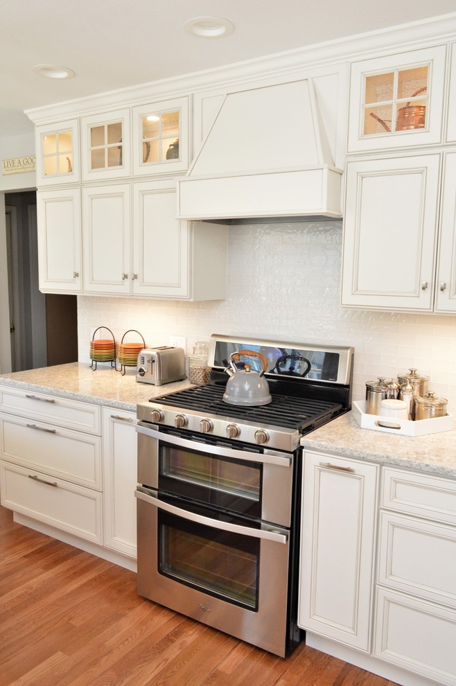 This is an example of a large traditional l-shaped open plan kitchen in Chicago with a farmhouse sink, recessed-panel cabinets, white cabinets, quartzite benchtops, white splashback, glass tile splashback, stainless steel appliances, medium hardwood floors, with island, brown floor and grey benchtop.