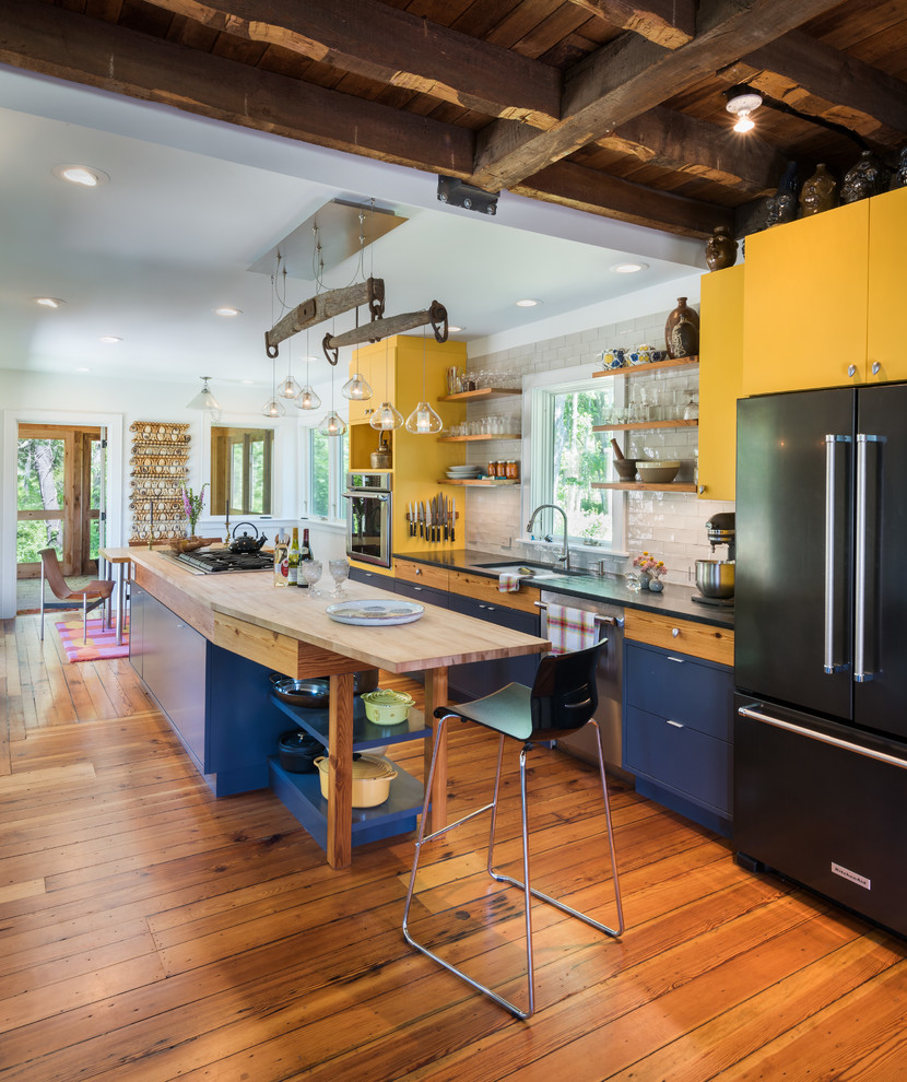 Photo of an eclectic galley eat-in kitchen in Providence with an undermount sink, flat-panel cabinets, yellow cabinets, white splashback, subway tile splashback, black appliances, medium hardwood floors, with island and black benchtop.