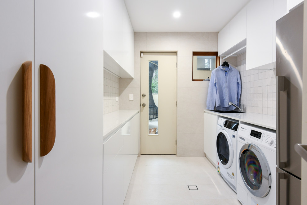 This is an example of a mid-sized modern galley dedicated laundry room in Sydney with a drop-in sink, flat-panel cabinets, white cabinets, quartz benchtops, beige splashback, ceramic splashback, beige walls, ceramic floors, a side-by-side washer and dryer, beige floor and white benchtop.