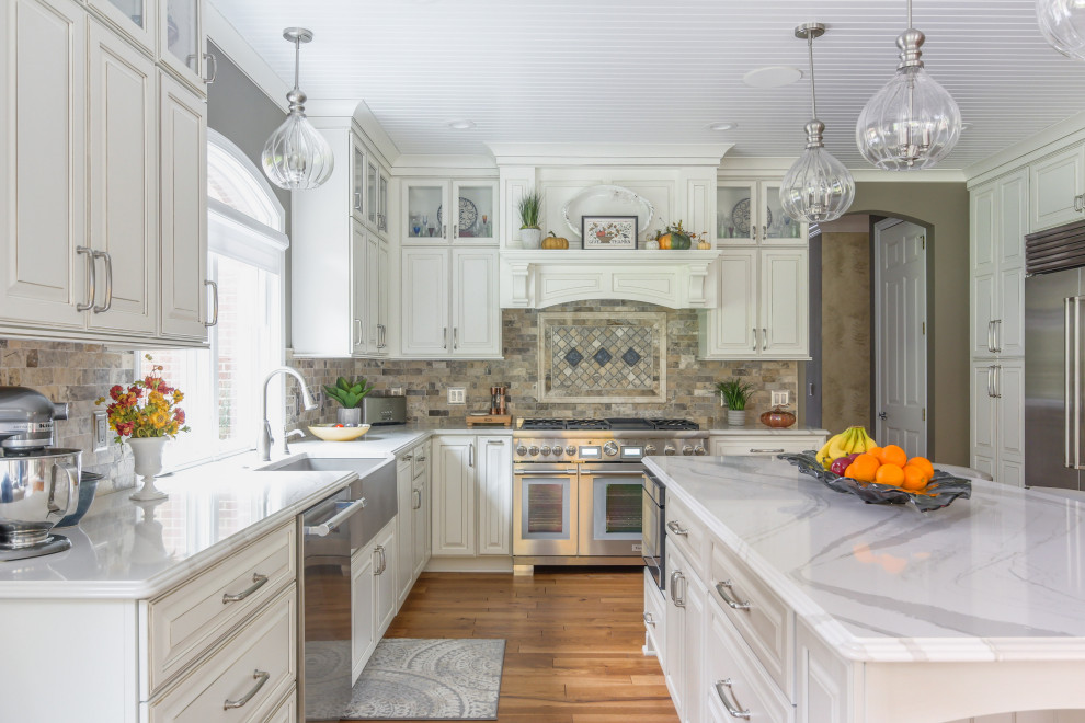 Large transitional l-shaped eat-in kitchen in Detroit with a farmhouse sink, raised-panel cabinets, white cabinets, quartz benchtops, beige splashback, travertine splashback, stainless steel appliances, medium hardwood floors, with island, brown floor and white benchtop.
