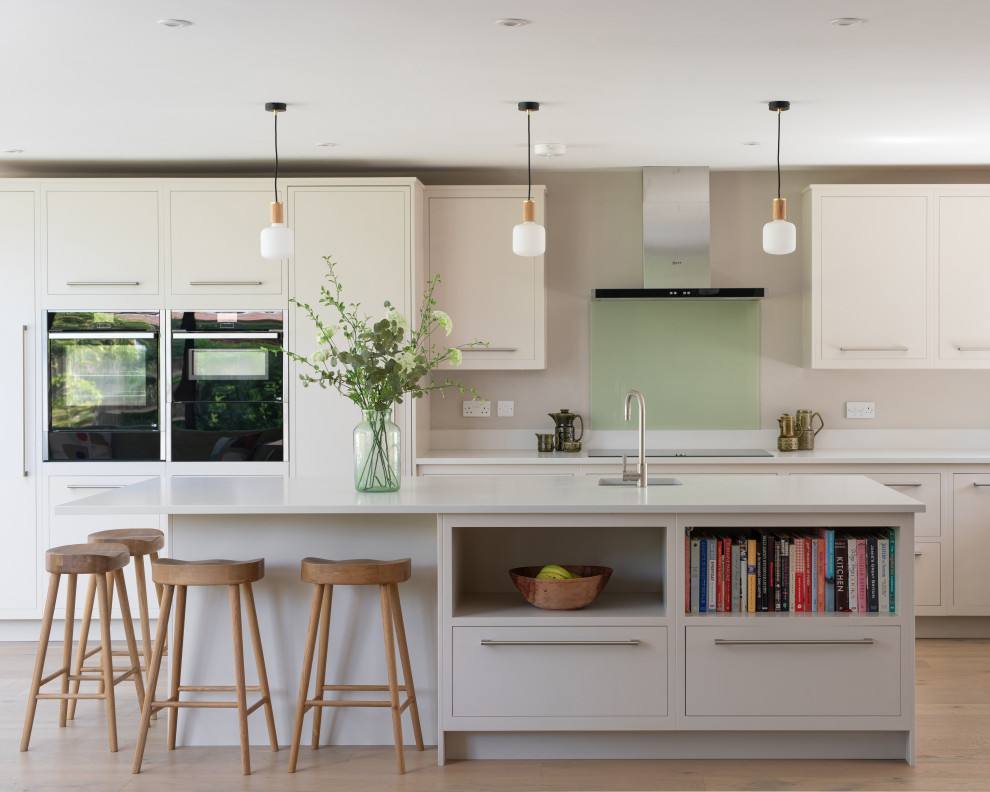 Design ideas for a medium sized contemporary grey and white l-shaped open plan kitchen in Sussex with a single-bowl sink, flat-panel cabinets, grey cabinets, quartz worktops, green splashback, glass sheet splashback, stainless steel appliances, light hardwood flooring, an island, brown floors and white worktops.