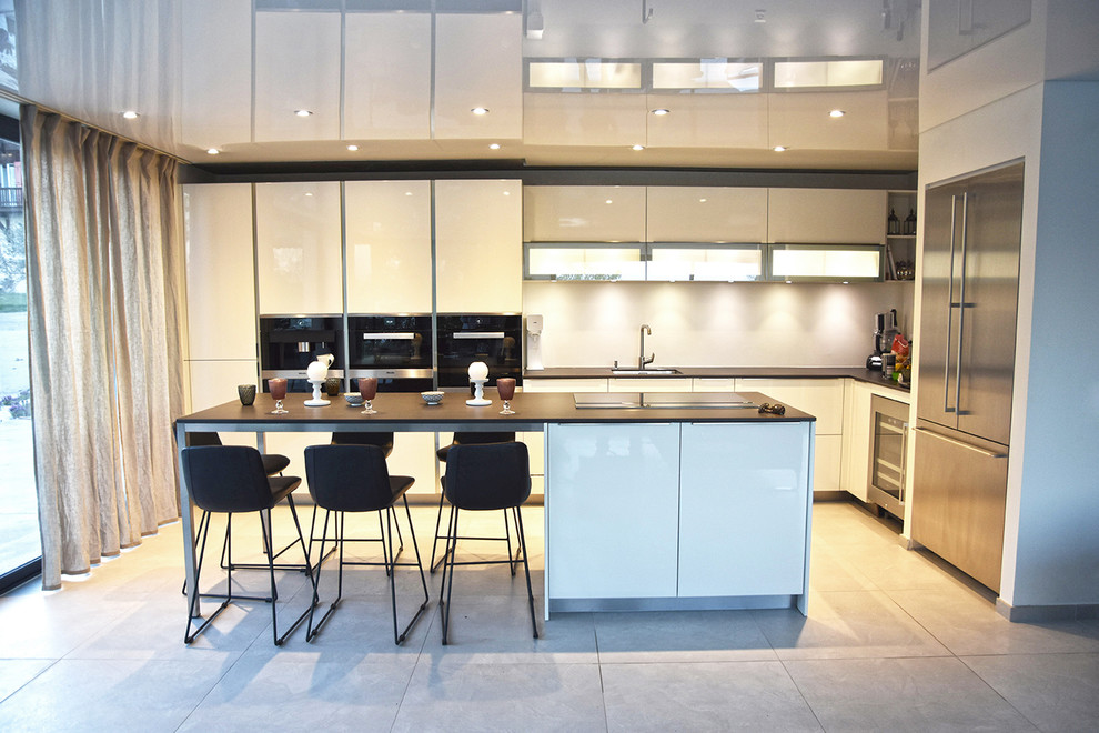 Photo of a large modern l-shaped open plan kitchen with an undermount sink, white cabinets, white splashback, with island, grey floor, beaded inset cabinets, limestone benchtops, glass sheet splashback, black appliances and ceramic floors.