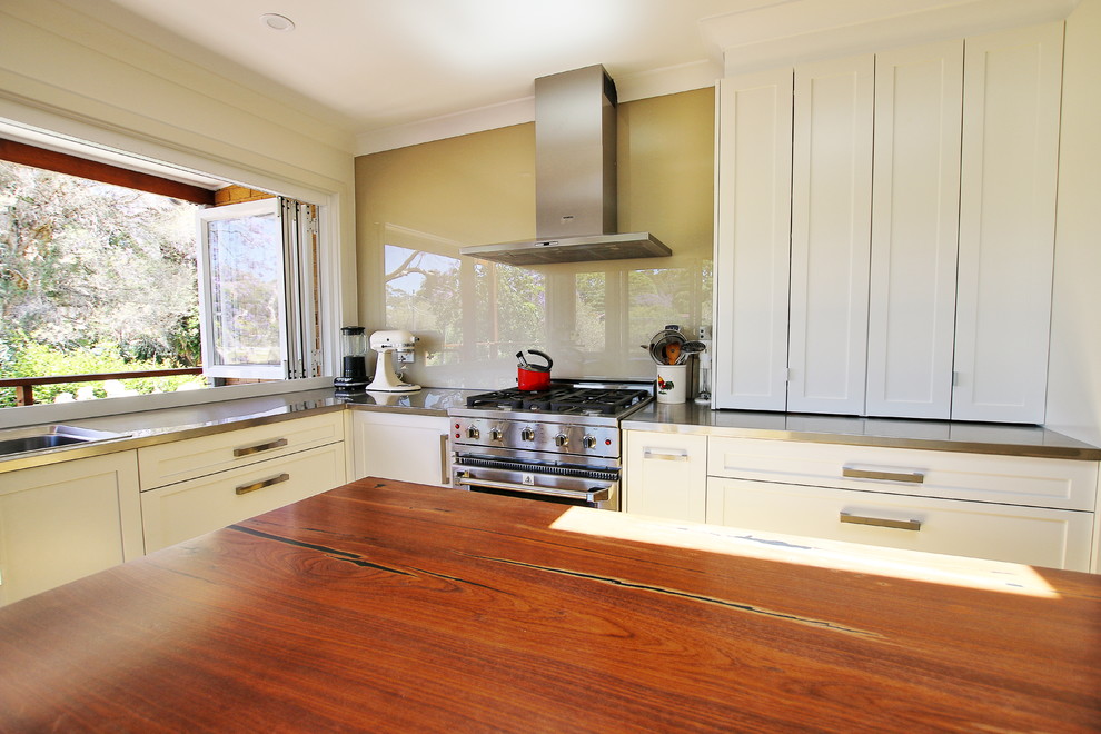Inspiration for a mid-sized transitional l-shaped kitchen pantry in Sydney with a double-bowl sink, shaker cabinets, white cabinets, wood benchtops, black splashback, glass sheet splashback, stainless steel appliances, light hardwood floors and with island.