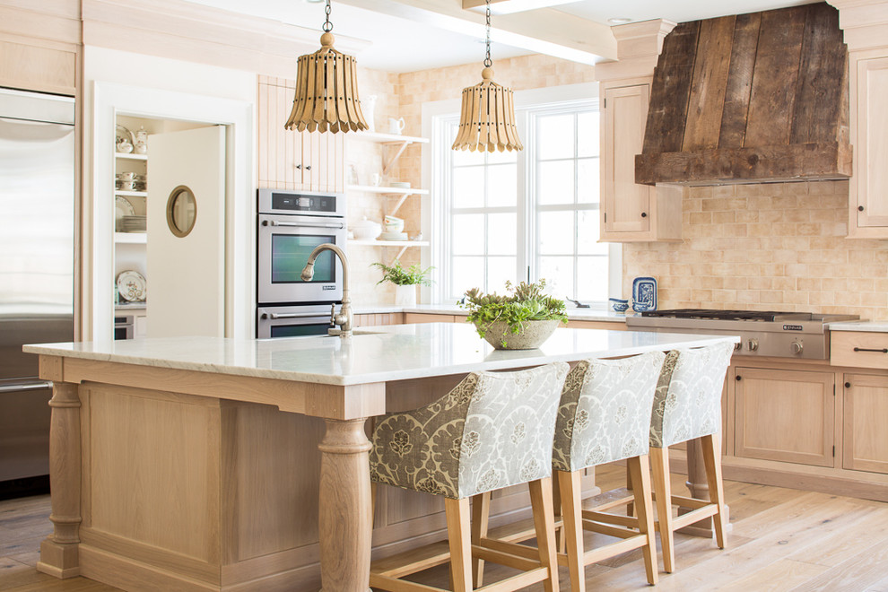 This is an example of a beach style l-shaped kitchen in Portland Maine with shaker cabinets, light wood cabinets, beige splashback, stainless steel appliances, light hardwood floors and with island.