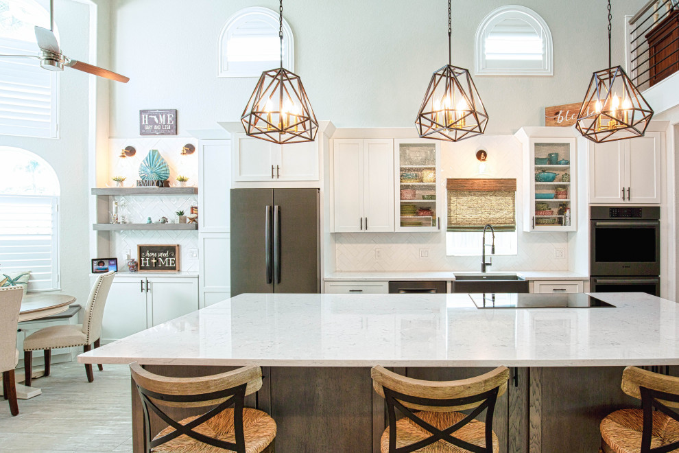 This is an example of a large beach style single-wall kitchen in Orlando with a farmhouse sink, shaker cabinets, distressed cabinets, quartz benchtops, white splashback, porcelain splashback, stainless steel appliances, porcelain floors, with island, beige floor and white benchtop.