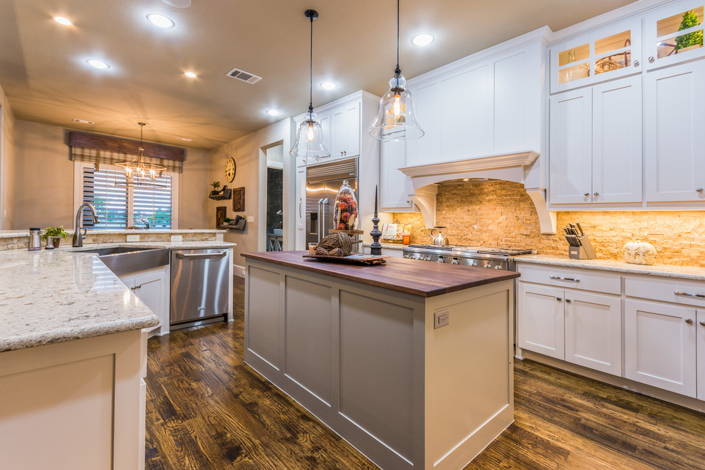 Inspiration for a large transitional l-shaped open plan kitchen in Dallas with a farmhouse sink, shaker cabinets, white cabinets, granite benchtops, brown splashback, stone tile splashback, stainless steel appliances, dark hardwood floors and multiple islands.