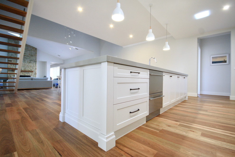 Large transitional l-shaped open plan kitchen in Sydney with an undermount sink, shaker cabinets, white cabinets, quartz benchtops, white splashback, subway tile splashback, stainless steel appliances, medium hardwood floors, with island, brown floor and grey benchtop.