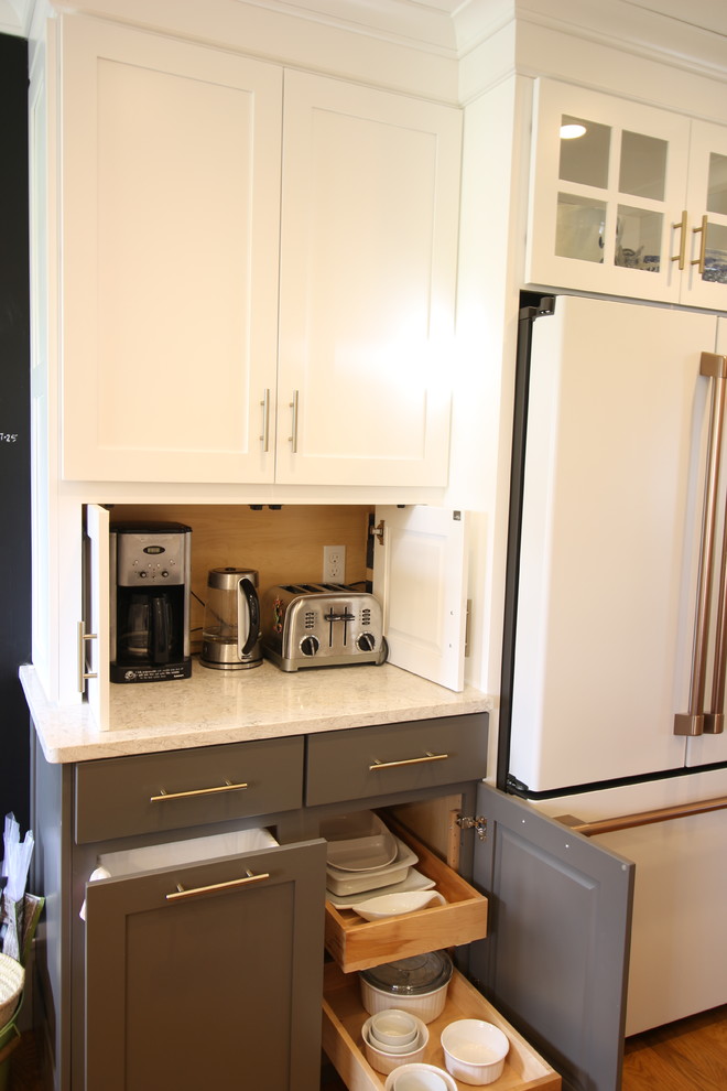 Example of a mid-sized transitional u-shaped medium tone wood floor and brown floor enclosed kitchen design with a farmhouse sink, shaker cabinets, white cabinets, quartz countertops, white backsplash, subway tile backsplash, white appliances, no island and beige countertops