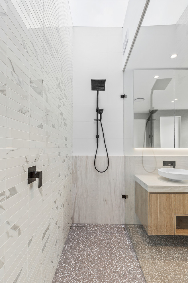 This is an example of a mid-sized modern master bathroom in Melbourne with flat-panel cabinets, light wood cabinets, a freestanding tub, an alcove shower, a wall-mount toilet, white tile, porcelain tile, white walls, terrazzo floors, a vessel sink, marble benchtops, grey floor and white benchtops.
