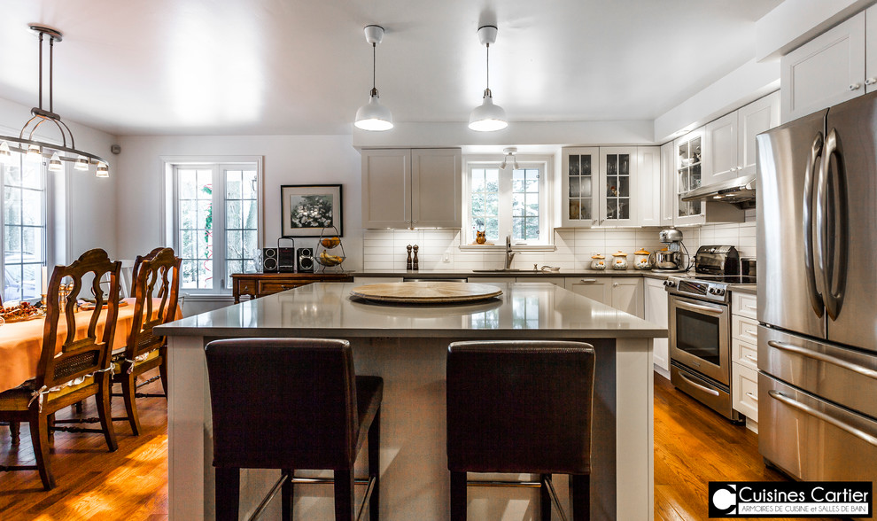 Eat-in kitchen - large transitional medium tone wood floor eat-in kitchen idea in Montreal with a double-bowl sink, shaker cabinets, white cabinets, quartzite countertops, white backsplash, ceramic backsplash, stainless steel appliances and an island