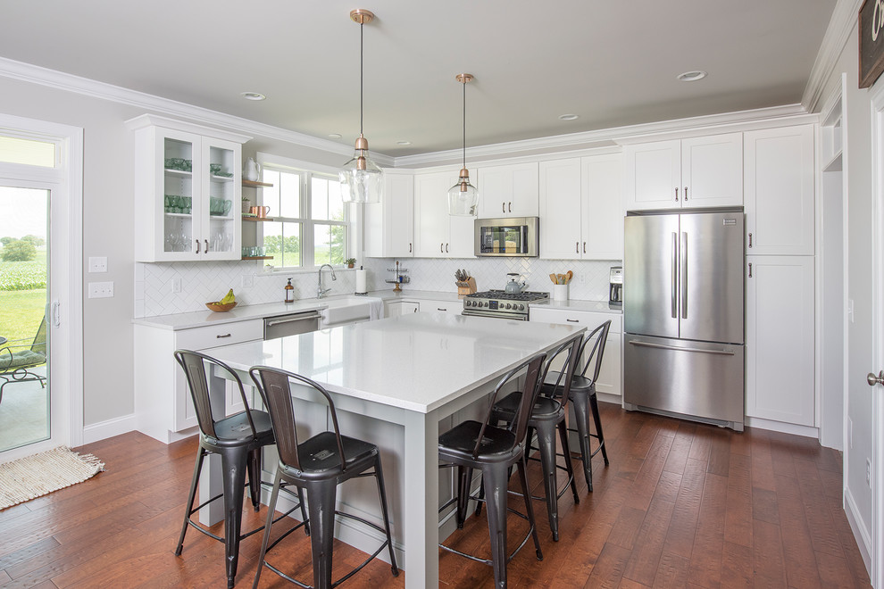 This is an example of a mid-sized transitional l-shaped open plan kitchen in Other with a farmhouse sink, shaker cabinets, white cabinets, quartzite benchtops, white splashback, subway tile splashback, stainless steel appliances, medium hardwood floors, with island and brown floor.
