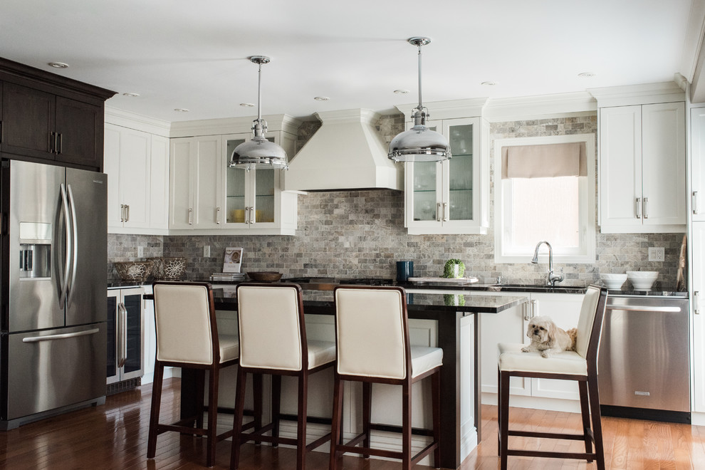 Photo of a transitional l-shaped kitchen in Toronto with an undermount sink, recessed-panel cabinets, white cabinets, granite benchtops, grey splashback, travertine splashback, stainless steel appliances, medium hardwood floors, with island, brown floor and black benchtop.