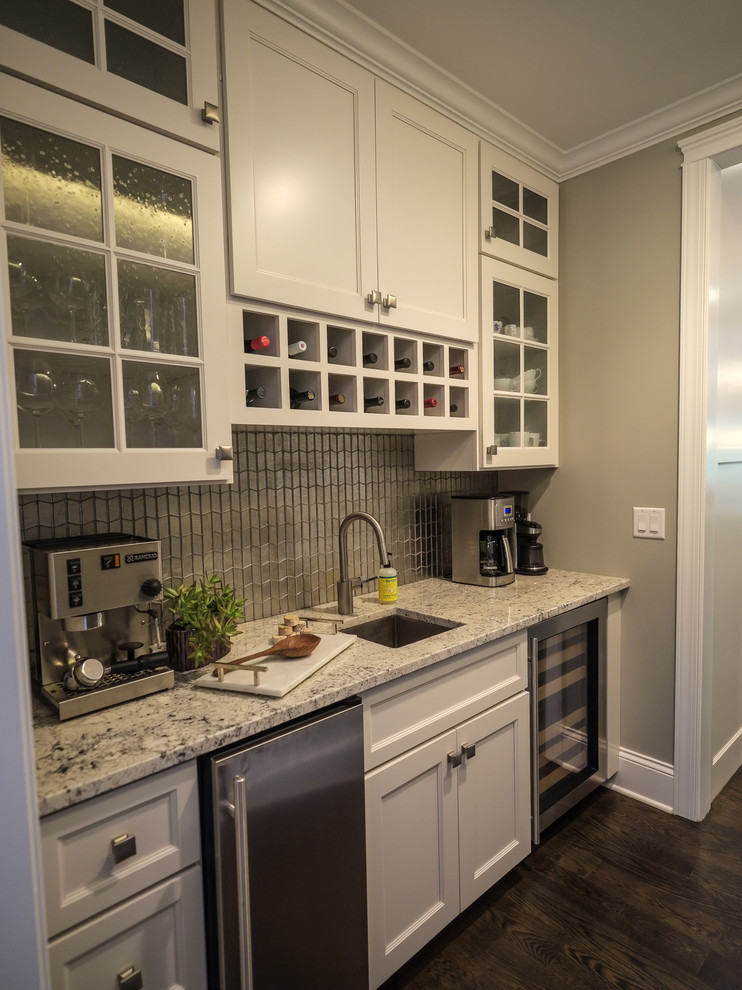 This is an example of a mid-sized transitional u-shaped wet bar in Chicago with an undermount sink, recessed-panel cabinets, white cabinets, granite benchtops, white splashback, subway tile splashback, dark hardwood floors, brown floor and white benchtop.