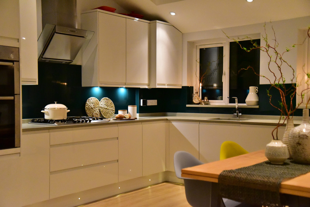 This is an example of a mid-sized contemporary u-shaped open plan kitchen in London with a single-bowl sink, flat-panel cabinets, white cabinets, solid surface benchtops, green splashback, glass sheet splashback, stainless steel appliances and laminate floors.