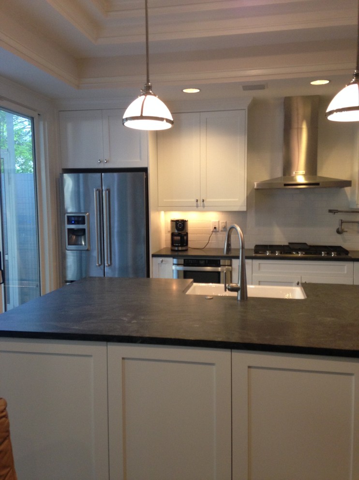 Example of a transitional l-shaped kitchen design in Atlanta with a farmhouse sink, shaker cabinets, white cabinets, granite countertops, white backsplash, ceramic backsplash, stainless steel appliances and an island