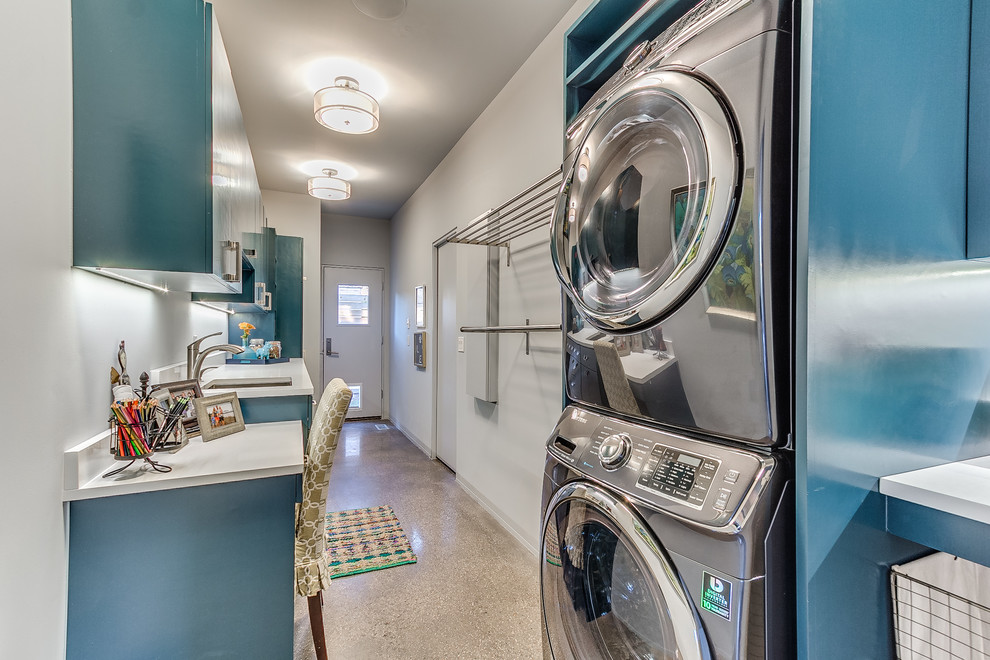Photo of a mid-sized contemporary single-wall utility room in Oklahoma City with an undermount sink, flat-panel cabinets, blue cabinets, quartz benchtops, white walls, concrete floors, a stacked washer and dryer and beige floor.